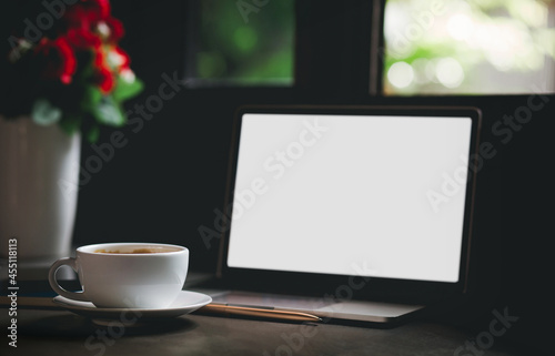 Focus on white coffee cup with computer laptop and vase with flower near window with sunlight shining through  Dark tone