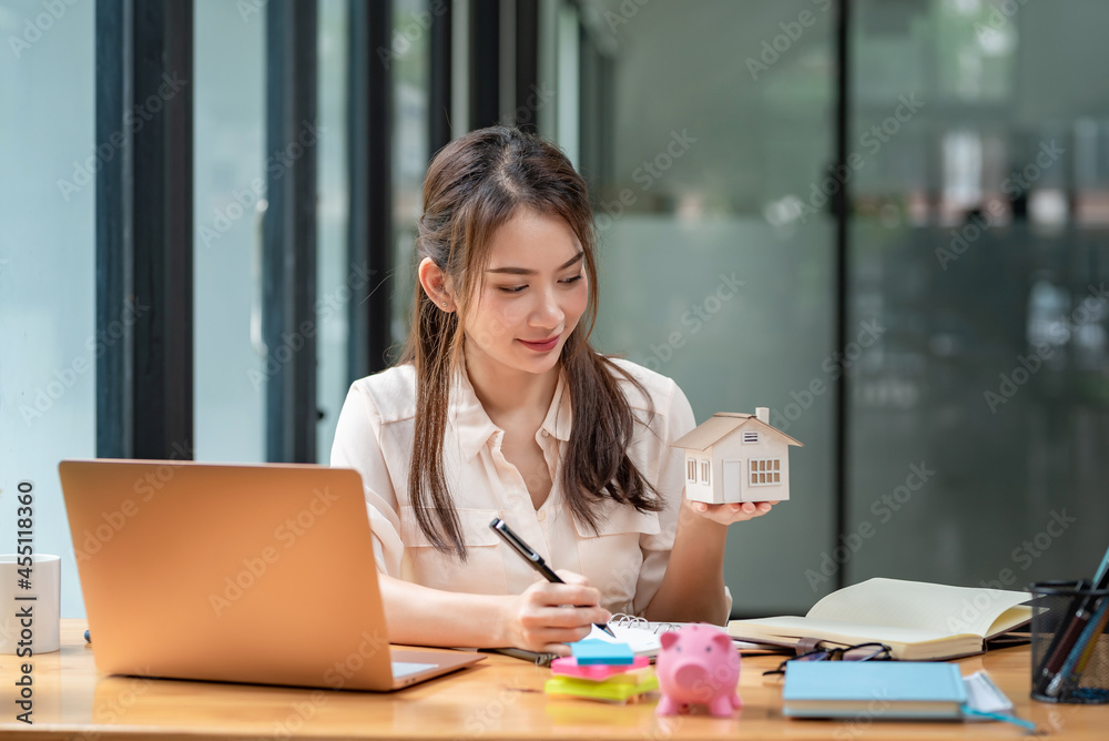 Young Asian Businesswoman or real estate agent holding a house example taking notes on a laptop with documents placed at the office. Young Asian Businesswoman or real estate agent holding a house example taking notes on a laptop with documents placed at the office.
