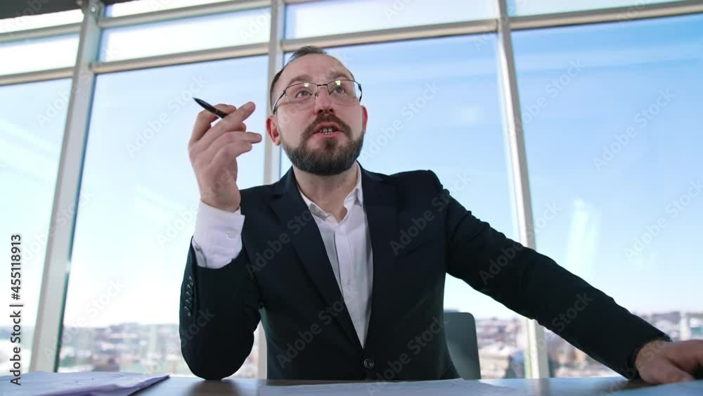 Portrait of a businessman sitting in office. Bearded entrepreneur in suit and glasses talking about business on window backdrop. Portrait of a businessman sitting in office. Bearded entrepreneur in suit and glasses talking about business on window backdrop.