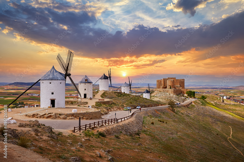 Wind mills and old castle in Consuegra, Toledo, Castilla La Mancha, Spain. Picturesque panorama landscape with road and view to ancient walls and windmills on blue sky with clouds. - obrazy, fototapety, plakaty 