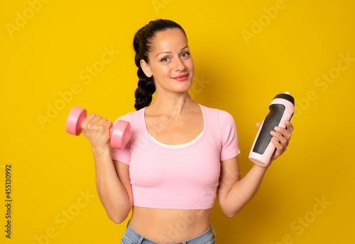 Young sportswoman holding a dumbbell and water isolated over yellow background.