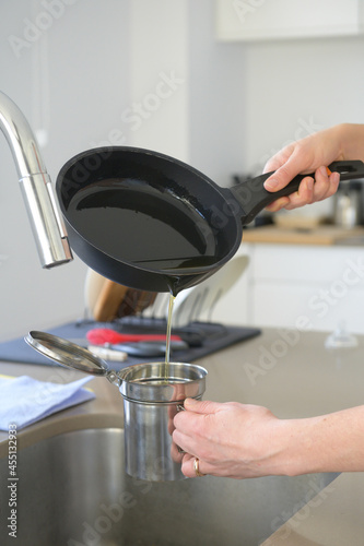 Woman pouring used cooking oil from an old frying pan into a metal cooking container in her kitchen.