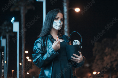 Chica joven de pelo castaño vestida de chaqueta disfrazada en la oscuridad para celebrar el dia de Halloween con su cesta de caramelos photo