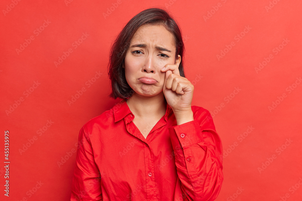 Upset gloomy Asian woman wipes tears after watching sad touching movie has heartbroken feelings feels discontent wears shirt poses against bright red background. Female model feels down depressed Upset gloomy Asian woman wipes tears after watching sad touching movie has heartbroken feelings feels discontent wears shirt poses against bright red background. Female model feels down depressed