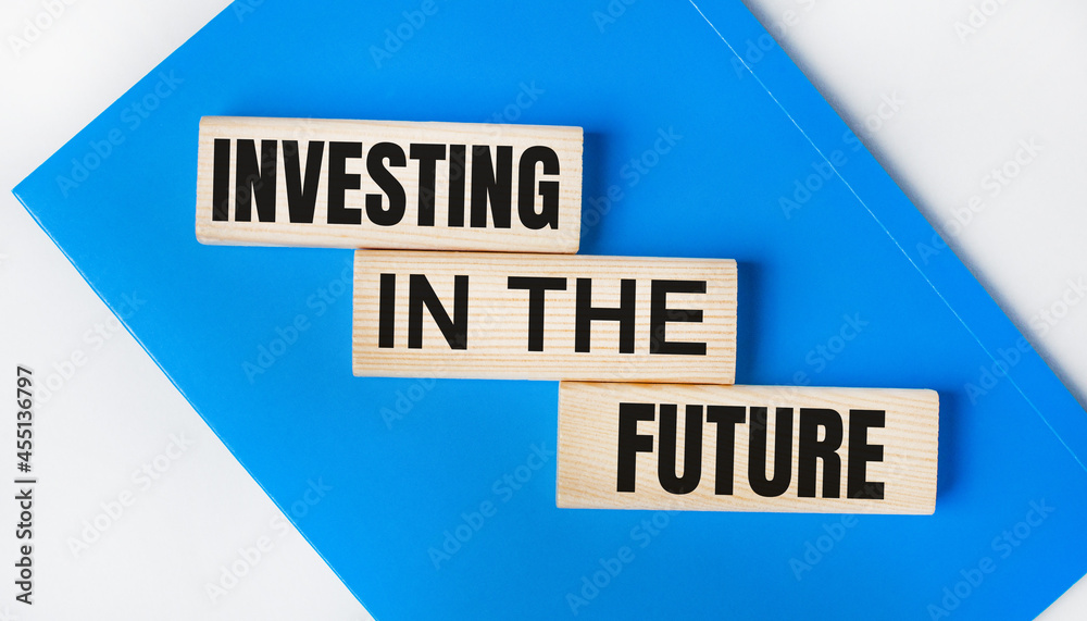 There is a blue notebook on a light gray background. Above are three wooden blocks with the words INVESTING IN THE FUTURE.