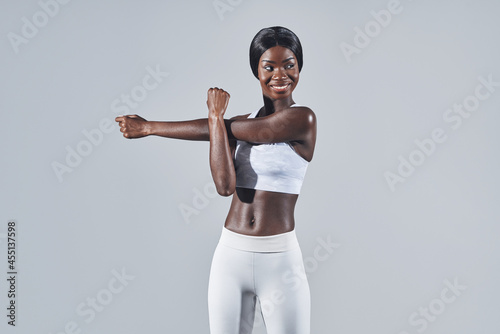 Beautiful young African woman in sports clothing stretching against gray background © gstockstudio