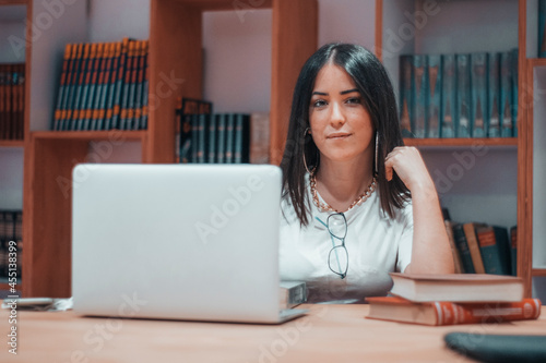 Muchacha joven estudiante revisando sus apuntes en el ordenador laptop en la biblioteca y sala de estudio junto a las estanterías y libros del instituto photo