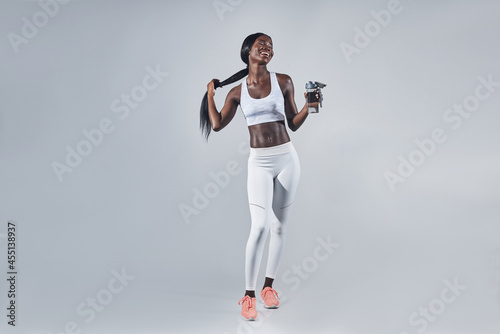 Fototapeta Naklejka Na Ścianę i Meble -  Happy young African woman in sports clothing holding bottle with water and adjusting hair