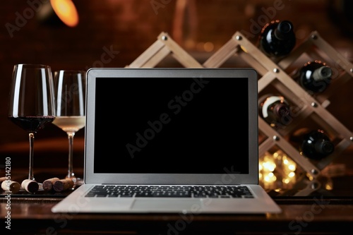 Empty blank screen laptop computer on table with wine bottle wine rack holder in the background. Buying wines online, home delivery concepts.