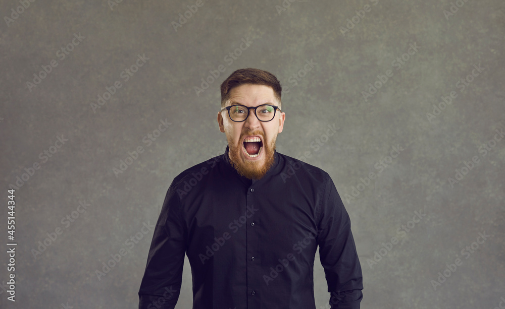 Crazy and furious Caucasian man in a rage shouts loudly and angrily looking into the camera. Man standing on a gray background feels stress and panic. Concept of frustration and negative emotions. Crazy and furious Caucasian man in a rage shouts loudly and angrily looking into the camera. Man standing on a gray background feels stress and panic. Concept of frustration and negative emotions.