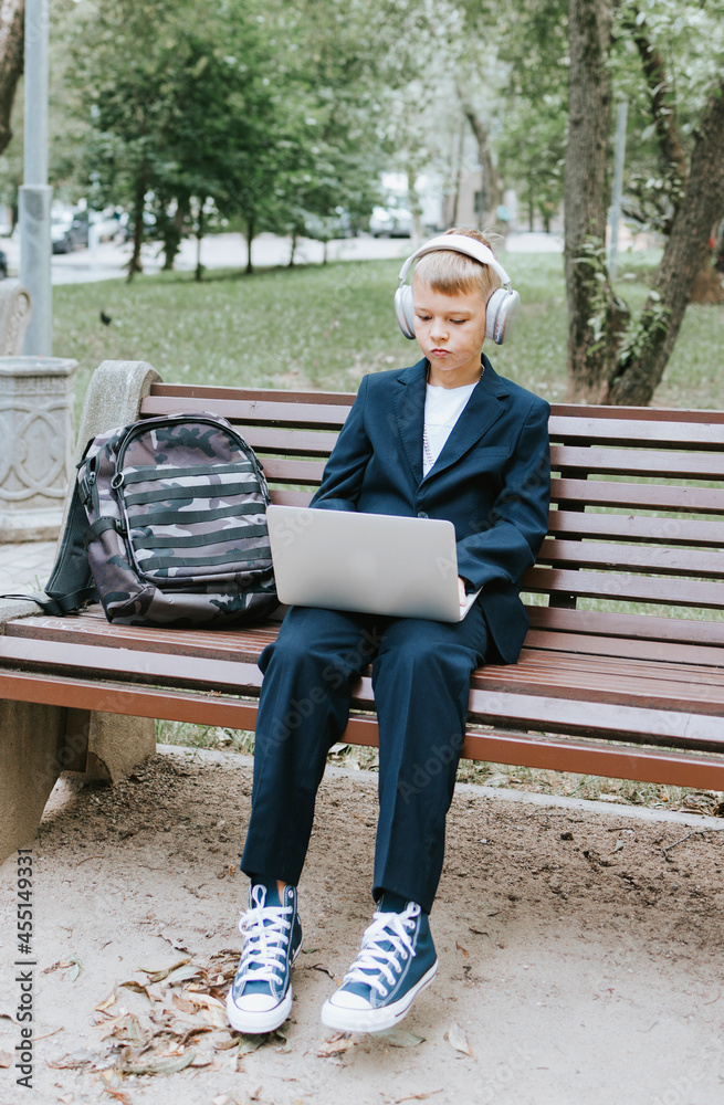 Fototapeta premium cute schoolboy with backpack in a school uniform with headphones and laptop is sitting on bench in the school yard, concept back to school and start of lessons