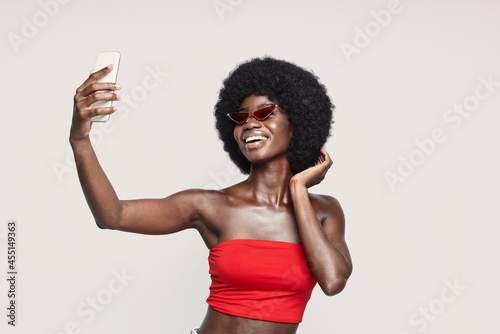 Beautiful young African woman making selfie by smart phone and smiling while standing against gray background