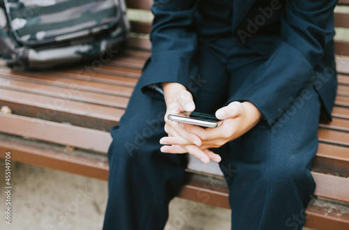 cute schoolboy with backpack in a school uniform with wireless headphones and using mobile phone is sitting on bench in the school yard, concept back to school and start of lessons