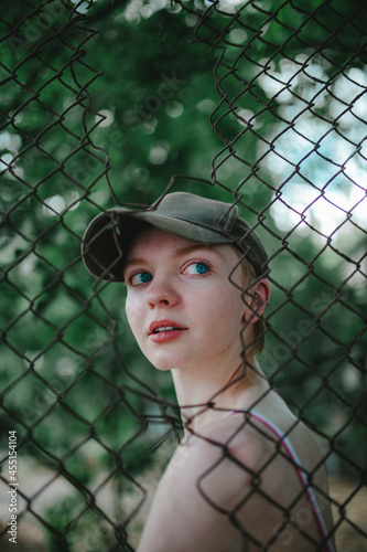 a girl with short hair in a baseball cap, looking through a mesh fence, through a mesh, bright blue eyes