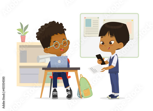 Smiling multinational primary classmates talking at classroom vector flat illustration. Happy schoolboy wearing school uniform use laptop, smartphone and paper sheet with task at lesson class interior