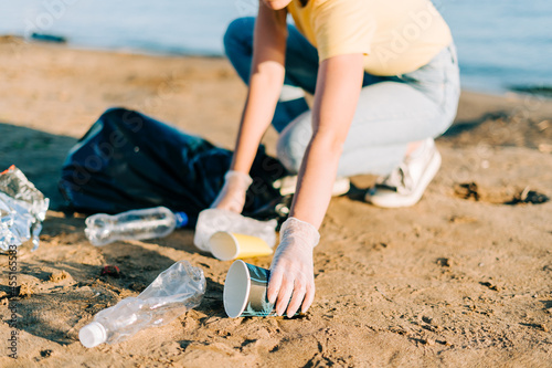 Young female volunteer hand satisfied with picking up trash, a plastic bottles and coffee cups, clean up beach with a sea. Woman collecting garbage. Environmental ecology pollution concept. Earth Day.