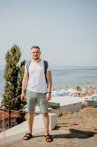  Young stylish man in white t-shirt and green jeans with a backpack walking and relaxing on a beach. Relaxed and cheerful. Outdoor photo of caucasian man near seaside, Summertime. travelling concept