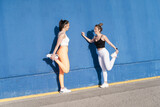Two female friends exercising together doing stretching workout over blue outdoors wall