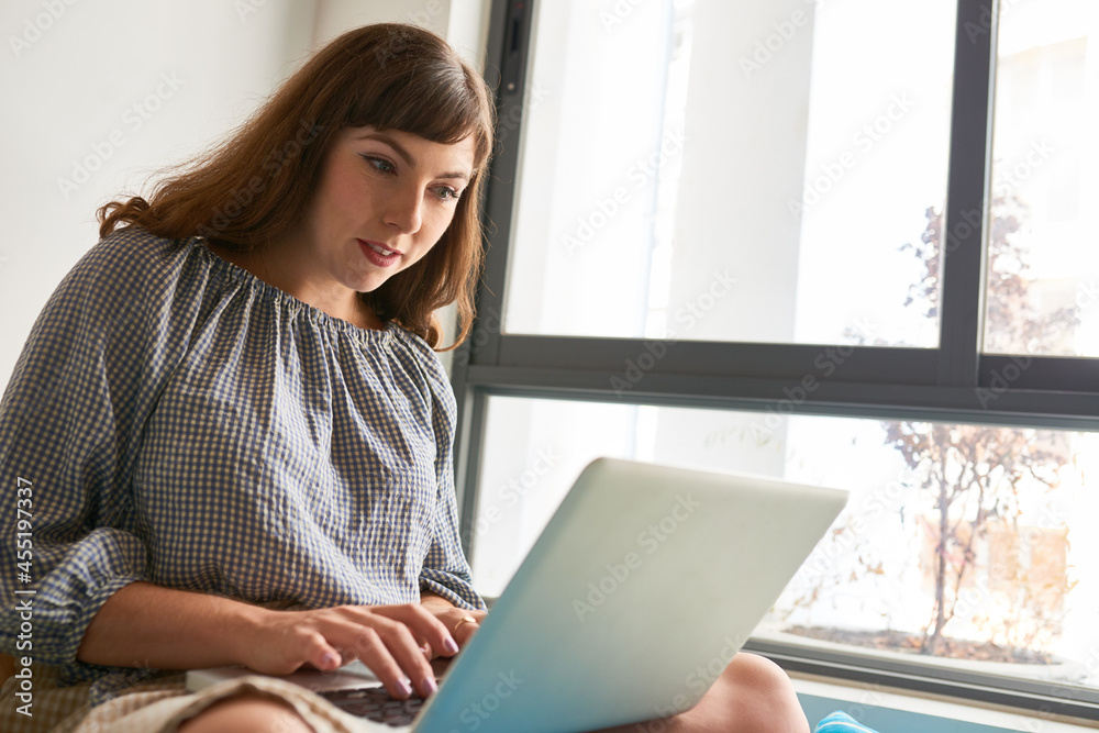 Positive woman sitting on window sill, coding or working with online documents Positive woman sitting on window sill, coding or working with online documents