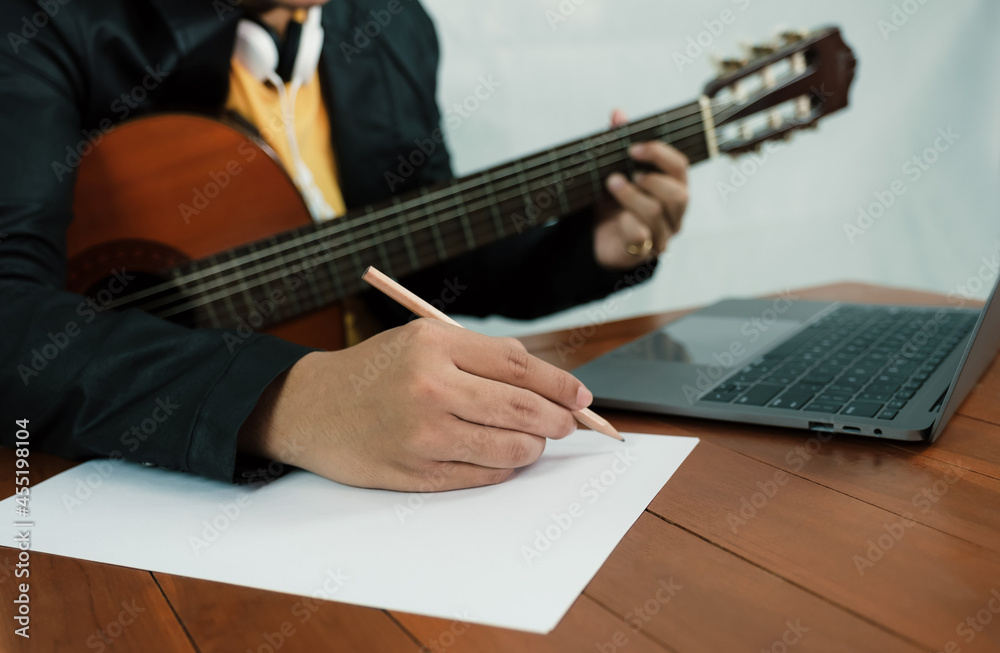 Fototapeta premium Young man musician hand holding a pencil to take notes of the music lesson, learning guitar online in the room with laptop