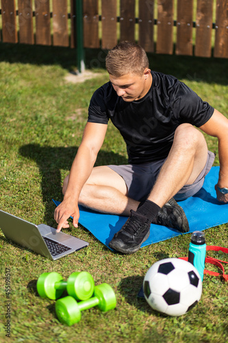 Close-up of a young man in a sports uniform is resting on lawn in garden, watches a movie and studies from a laptop , a social network. Student loafing around and putting off work and study
