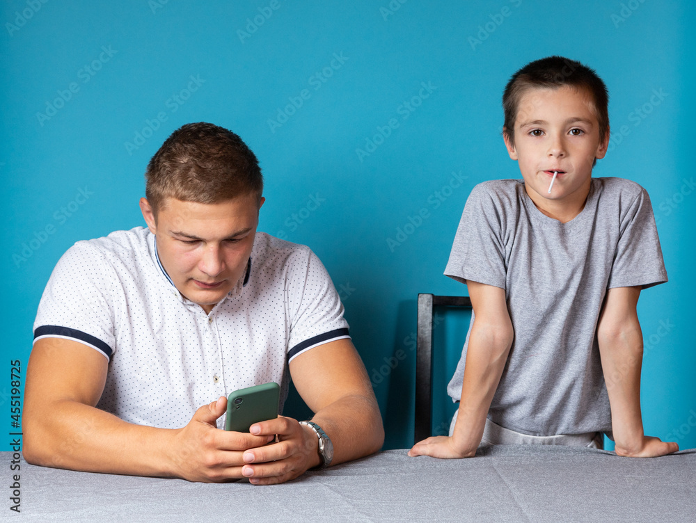 Home lessons with the child, dad is distracted by the phone while the son is bored and waiting for his attention Home lessons with the child, dad is distracted by the phone while the son is bored and waiting for his attention