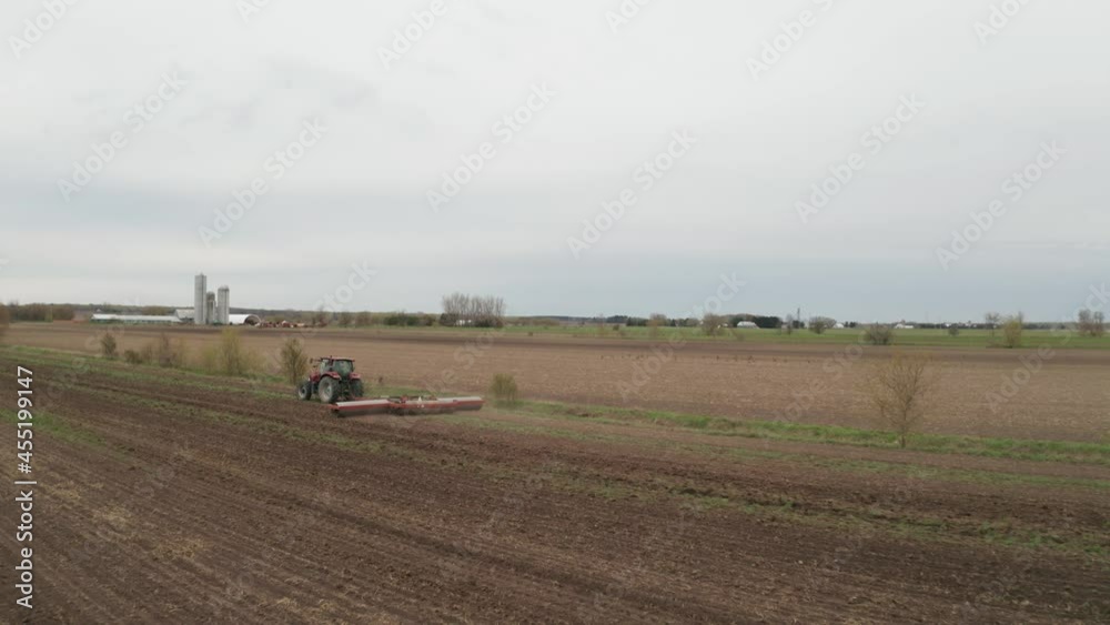 Farmer working in field driving tractor towing machinery to plow, till, disc a field for planting. Barn and silos in distance. Aerial view from drone flying over agricultural landscape and farm. Farmer working in field driving tractor towing machinery to plow, till, disc a field for planting. Barn and silos in distance. Aerial view from drone flying over agricultural landscape and farm.