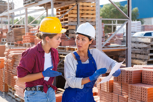 Two female storekeepers working in a building materials store are discussing important work issues in the warehouse, holding ..an estimate in their hands