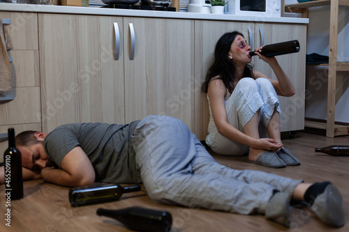 Bruised victim drinking alcohol after domestic physical abuse while husband with bottles laying on floor. Crying unhappy woman feeling miserable because of social problems and violence photo