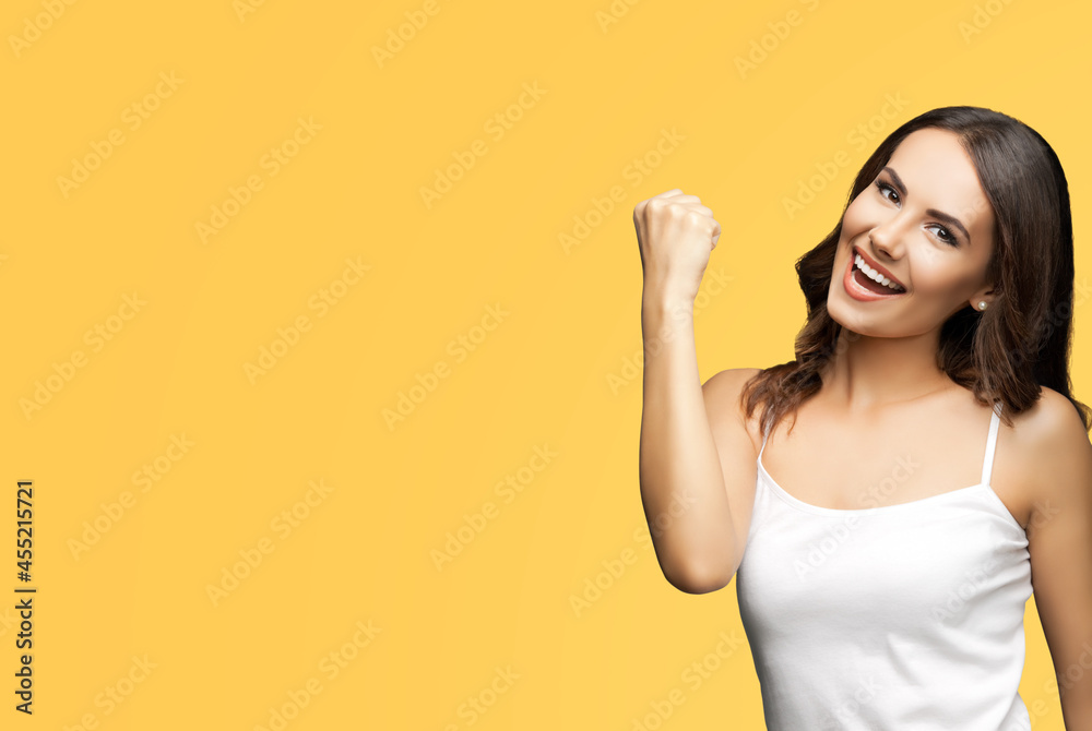 Photo of happy gesturing beautiful woman with open mouth raising hand up with clenching fist, in white smart casual clothing, isolated over yellow color background. Excited brunette girl at studio. Photo of happy gesturing beautiful woman with open mouth raising hand up with clenching fist, in white smart casual clothing, isolated over yellow color background. Excited brunette girl at studio.