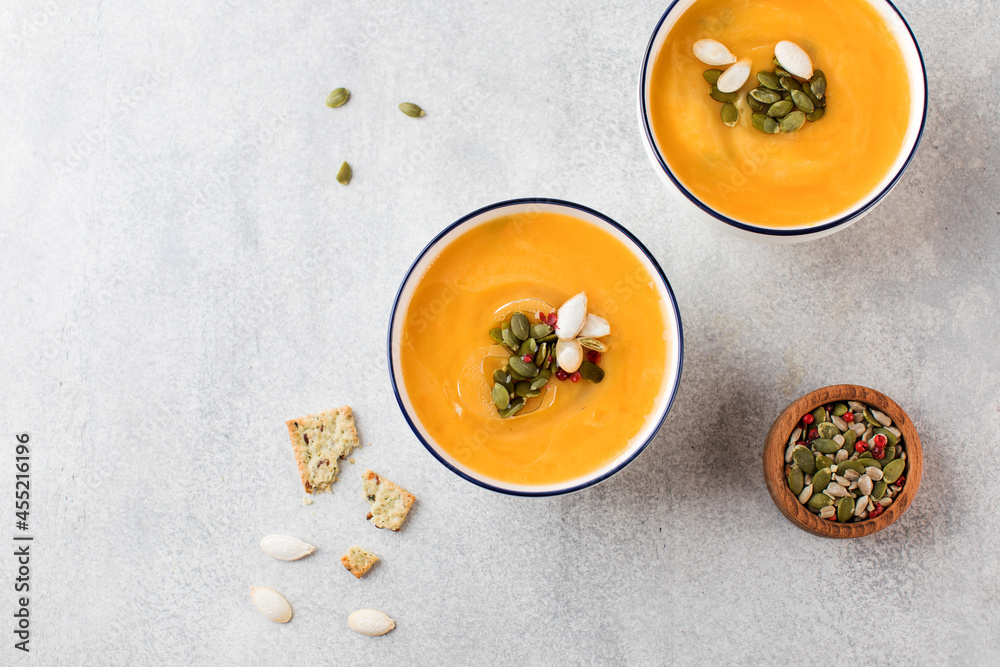 Two bowls with pumpkin cream soup and pumpkin seeds on a gray background. Top view. Autumn recipes. Two bowls with pumpkin cream soup and pumpkin seeds on a gray background. Top view. Autumn recipes.