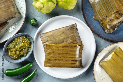 Tamales oaxaquenos, traditional dish of the cuisine of Mexico, various stuffings wrapped in green leaves, overhead flat lay shot. Hispanic food. With chili peppers, tomatillos, and salsa verde photo