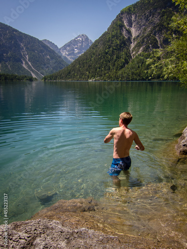 man in swimming trousers walking in to a nice lake with mountains in the background and is throwing a stone