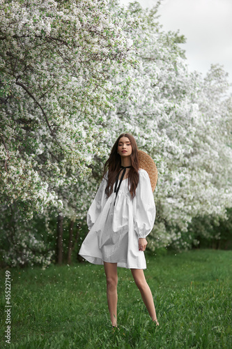 Young woman in white dress with a straw hat walks through blooming spring garden park. Spring has come, romantic mood