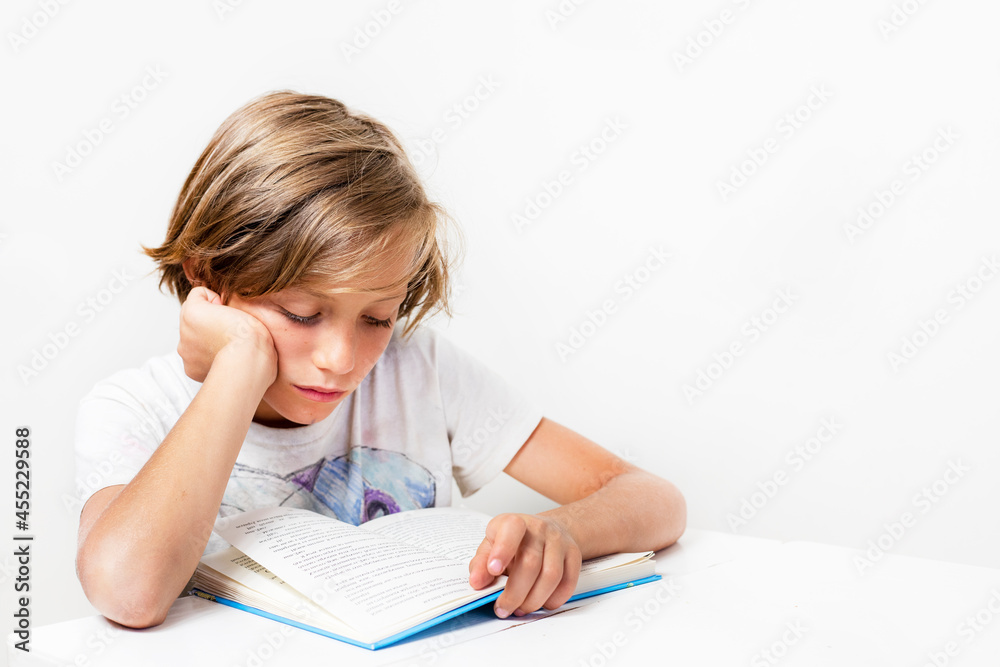 serious boy reading an interesting book at a desk against a background of a white wall, back to school, Authenticity serious boy reading an interesting book at a desk against a background of a white wall, back to school, Authenticity