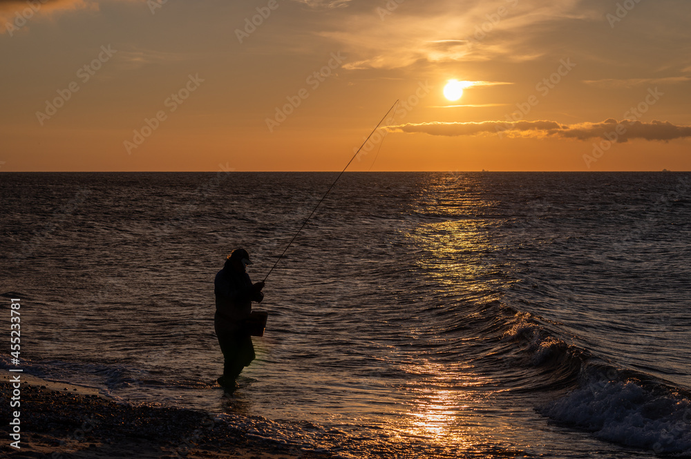 Angler steht mit seiner Angel am Meer im Abendrot bei Sonnenuntergang und angelt.