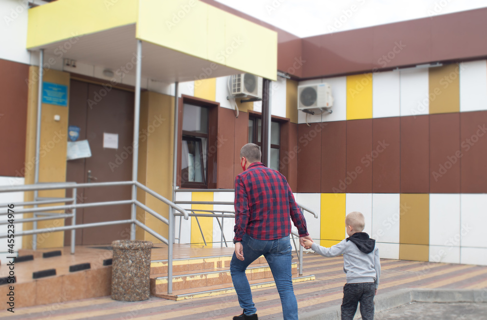 Fototapeta premium Dad takes his son to the doctor. Against the backdrop of a standard hospital building, father and son prepare for an examination. Concept of a single parent preparing a child for school or kindergarte