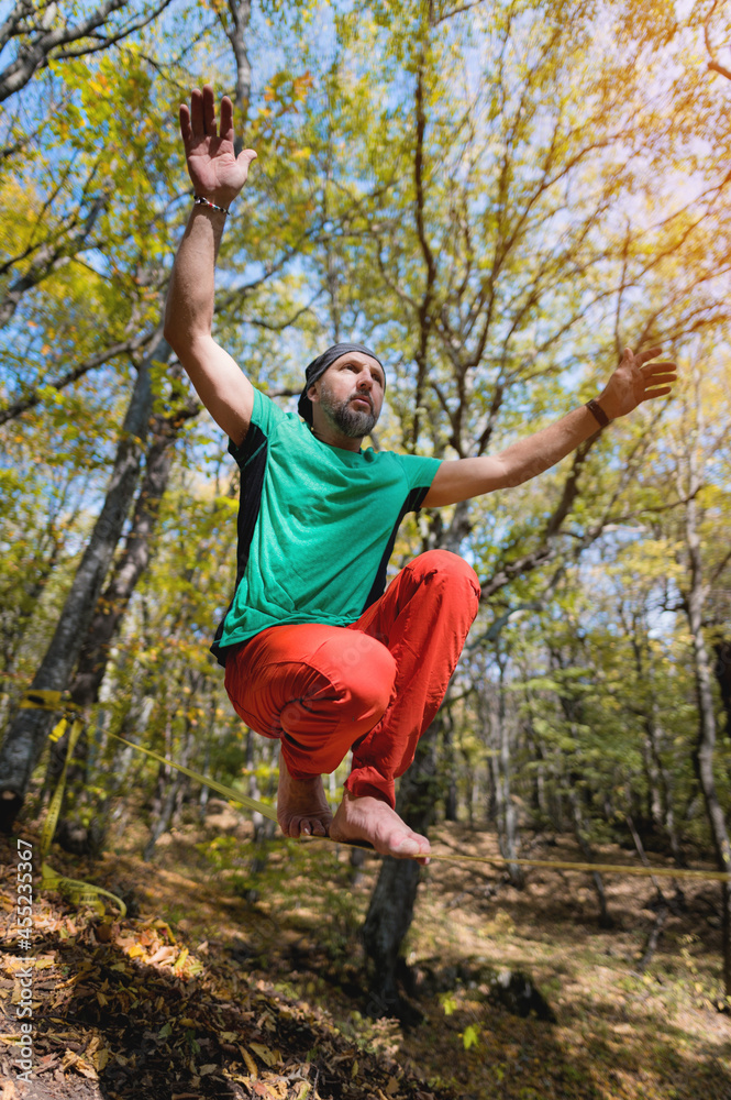 Mature male athlete doing a sitting trick while standing on the slackline. Sports activities to develop balance in the autumn outdoor forest Mature male athlete doing a sitting trick while standing on the slackline. Sports activities to develop balance in the autumn outdoor forest