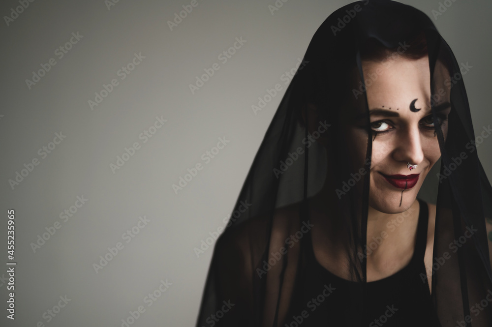 Redhead woman with a black veil on her head in a dark studio. A girl with an earring in her nose smiles slyly. Witch Makeup. Halloween costume. Redhead woman with a black veil on her head in a dark studio. A girl with an earring in her nose smiles slyly. Witch Makeup. Halloween costume.
