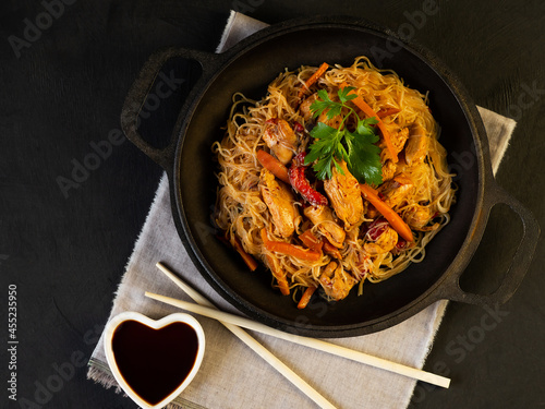 Classic wok noodles with chicken and vegetables in soy sauce, served in a cast iron pan on a black background with chopsticks.