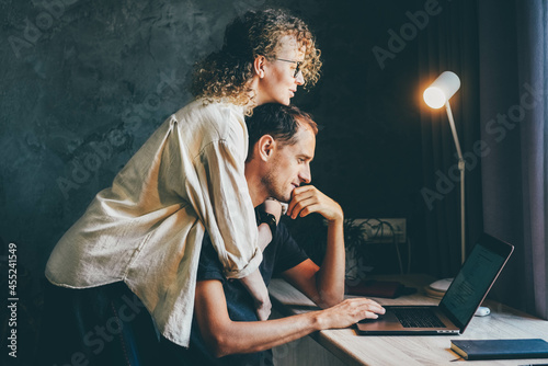 Curly haired woman hugs husband and looks at laptop screen working together on startup project at long desk near large window in home office.