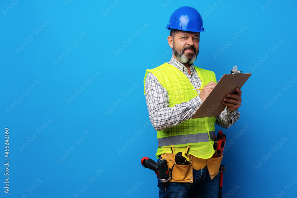 Male builder or handyman in uniform holding clipboard against blue background Male builder or handyman in uniform holding clipboard against blue background