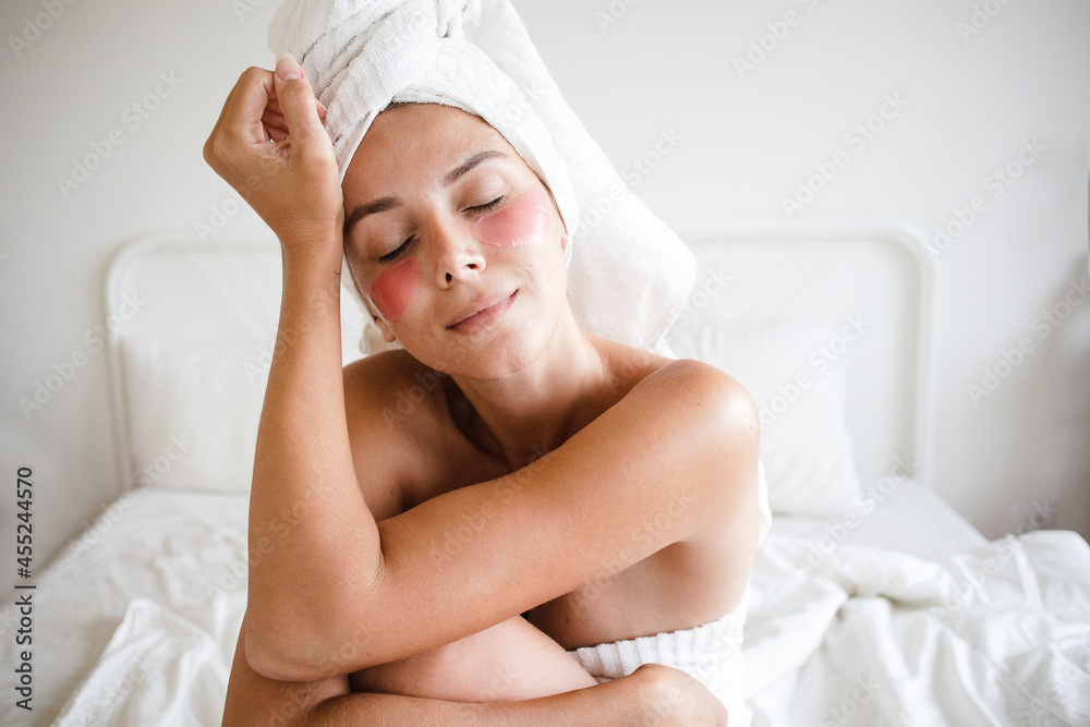 young white woman no makeup towel on head sitting on the bed in love with herself with pink under-eye patches  dark circles, puffiness and lines smiling softly and with eyes closed self care routine