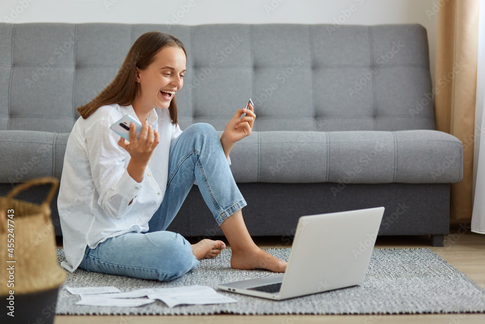 Portrait of happy woman wearing white shirt and jeans sitting on floor and looking at laptop screen, holding phone and card, checking bank account, winning lottery, crediting a large amount of money.
