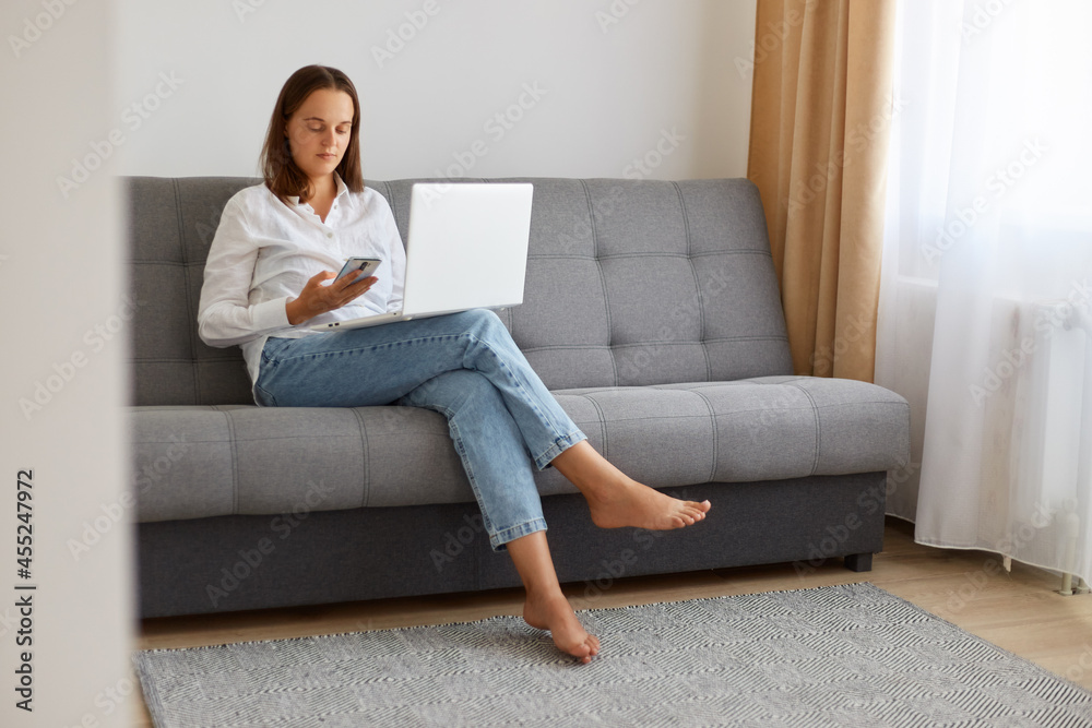 Indoor shot of calm confident female wearing white shirt and jeans sitting on sofa with laptop on legs, freelancer using phone while having break, posing in light living room. Indoor shot of calm confident female wearing white shirt and jeans sitting on sofa with laptop on legs, freelancer using phone while having break, posing in light living room.