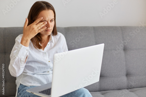 Photos Portrait of unhappy tired female wearing white shirt and jeans sitting on sofa at home, holding notebook and looking at computer screen, working long hours, feels hurt in eyes, rubbers eye