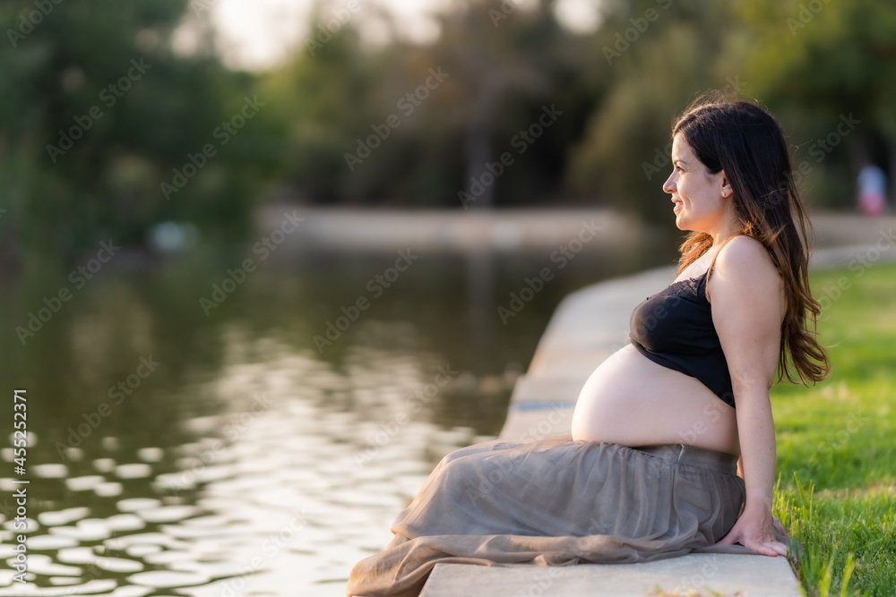 Pregnant woman contemplating the river of a park while sitting during sunset