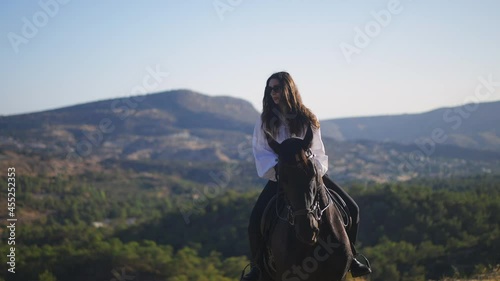 Portrait of gorgeous young woman sitting on brown horse in sunny mountains. Caucasian brunette horsewoman rider and graceful animal outdoors. Lifestyle and horse riding concept