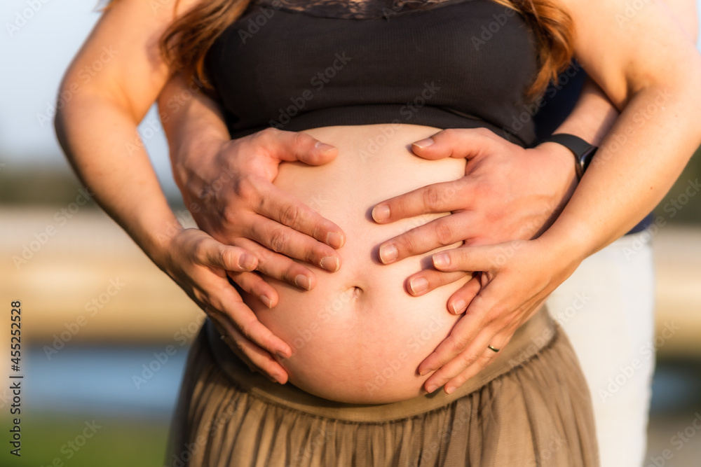 Close-up view of four hands on the abdomen of a pregnant adult woman standing outdoors Close-up view of four hands on the abdomen of a pregnant adult woman standing outdoors