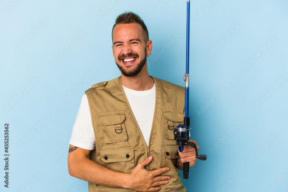 Young caucasian fisherman with tattoos holding rod isolated on blue background laughing and having fun. Young caucasian fisherman with tattoos holding rod isolated on blue background laughing and having fun.